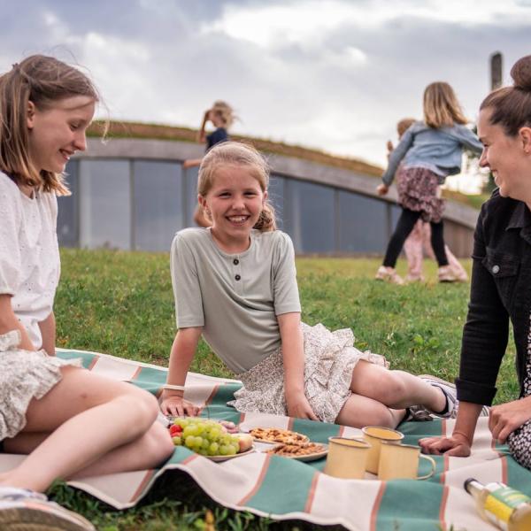 Eine Mutter und ihre beiden Töchter sitzen auf einer Decke und essen Snacks. Hinter ihnen befindet sich das Besucherzentrum, das in einen Erdwall gebaut ist.